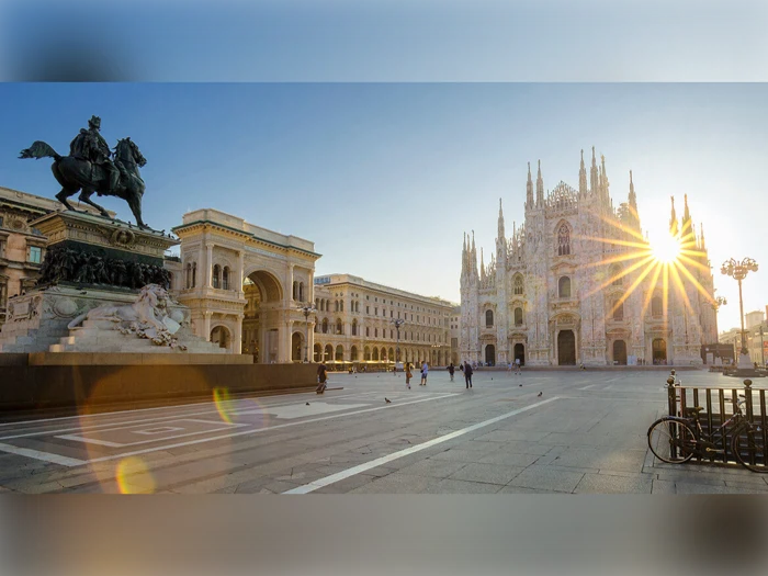 En solbeskinnet Piazza del Duomo i Milano med Milanos domkirke, en statue af en mand på hesteryg og et par mennesker, der går på pladsen.