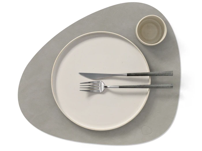 Minimalistisk borddækning med en rund beige tallerken, en lille beige skål, gaffel og kniv på en grå, asymmetrisk dækkeserviet.