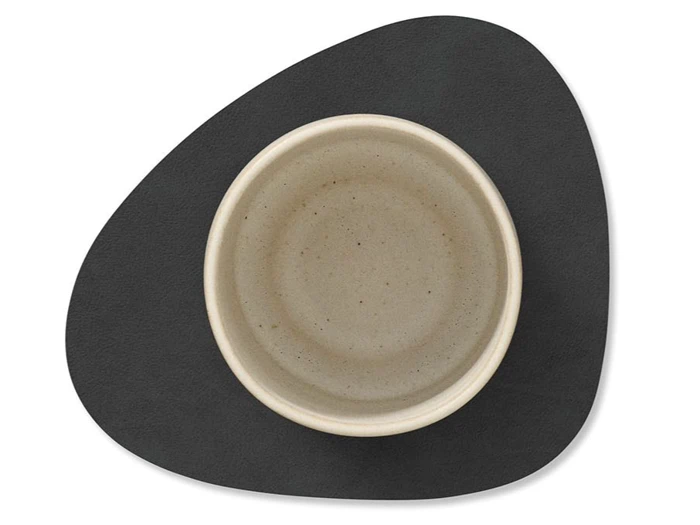 En rund, beige keramikskål står midt på en sort, uregelmæssigt formet dækkeserviet mod en hvid baggrund.