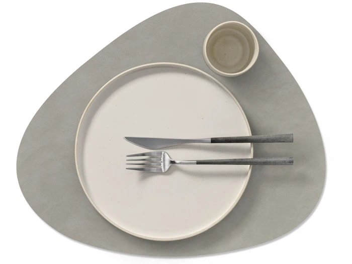 Minimalistisk borddækning med en rund tallerken, gaffel og kniv på kryds og tværs, en lille kop, alt sammen på en uregelmæssigt formet grå dækkeserviet.