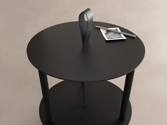 Rundt sort sidebord med en struktureret overflade, en håndtagsløkke på toppen og et lille foto og en kuglepen placeret på overfladen. Baggrunden er en neutral beige farve.