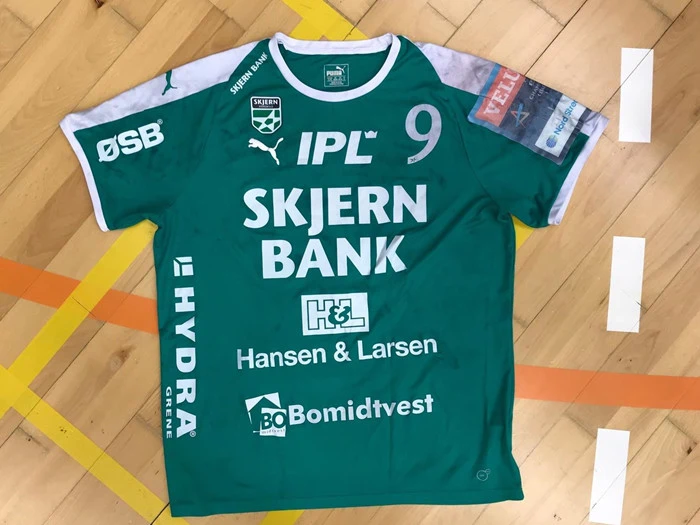 En grøn sportstrøje med hvid skrift, nummer 9 og flere sponsorer, bl.a. SKJERN BANK, ligger på et trægulv i en gymnastiksal med gule og orange banelinjer.
