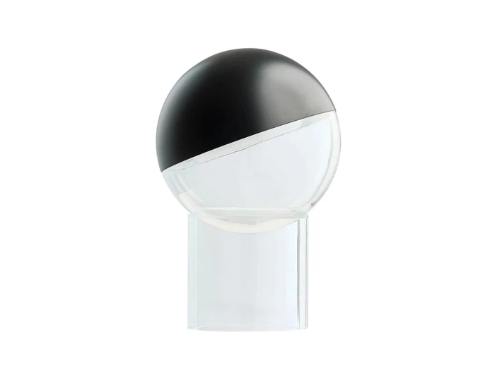 En moderne bordlampe med en rund kuppel, der er halvt sort og halvt gennemsigtig, og som står på en gennemsigtig, cylindrisk fod, set på en hvid baggrund.