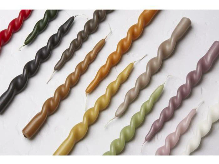 Assorterede snoede lys i forskellige farver, herunder rød, grøn, sort, gul, brun, lilla, hvid og orange, arrangeret diagonalt på en hvid struktureret overflade.