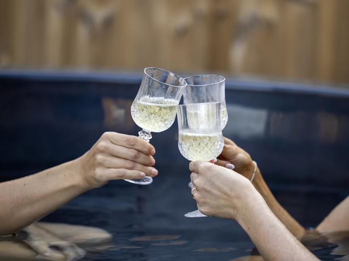 Tre hænder klirrer med vinglas fyldt med hvidvin over mørkt vand, muligvis mens de slapper af i et spabad eller en pool.