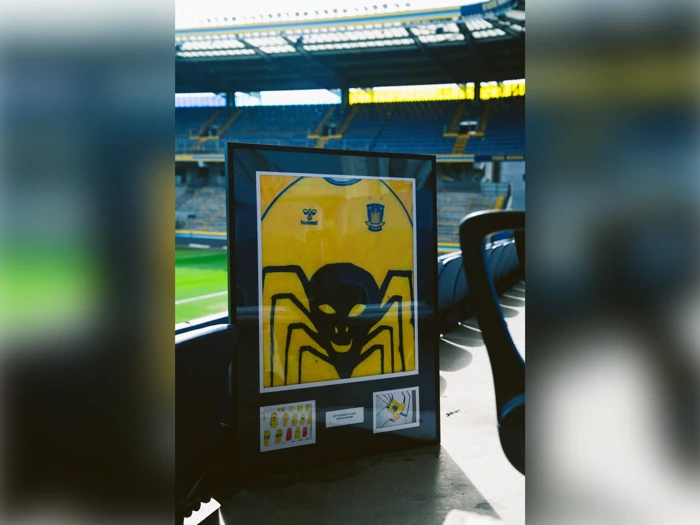 Indrammet gul fodboldtrøje med en sort edderkopgrafik på en baggrund af et fodboldstadion.