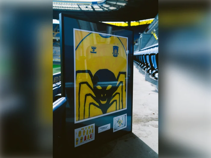 En indrammet gul fodboldtrøje med et stort sort edderkoppemotiv vises på et stadionsæde med tomme rækker og banen synlig i baggrunden.