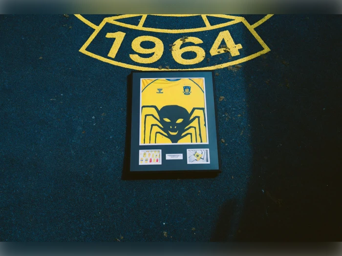 En indrammet gul sportstrøje med et stort sort edderkoppemotiv er placeret på mørk jord under en fed gul 1964-markering.