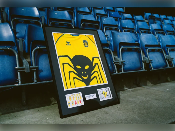 En indrammet gul fodboldtrøje med et stort sort edderkoppemotiv er udstillet på et stadionsæde med tomme blå sæder i baggrunden.