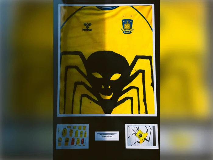 En indrammet gul fodboldtrøje med et stort sort edderkoppedesign, Brøndby IF-logo og tre små designskitser nedenunder er udstillet.