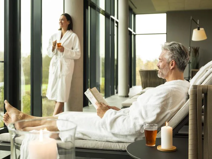 En mand slapper af i en lænestol og læser en bog med te og et stearinlys i nærheden, mens en kvinde står ved vinduet, begge i hvide badekåber, i et lyst, moderne spa-område.