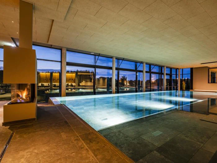 Moderne indendørs swimmingpool med store vinduer med udsigt til et udendørs område, stemningsfuld belysning og en tændt pejs til venstre.