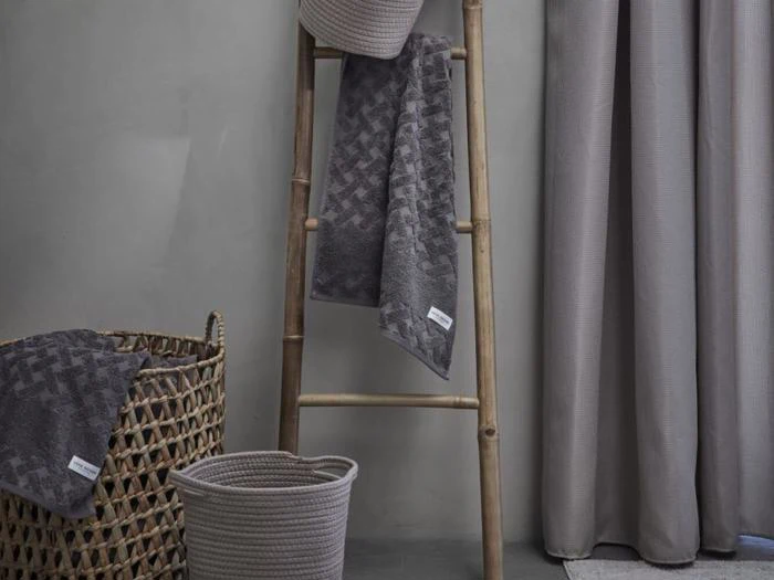 En bambusstige holder grå håndklæder ved siden af en flettet kurv og en stofspand; grå gardiner hænger i baggrunden.