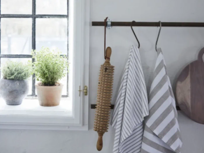 To stribede viskestykker og en kagerulle i træ hænger på en stang; to potteplanter står i vindueskarmen ved siden af.