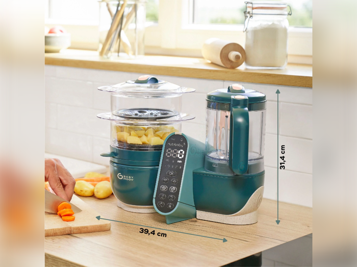 Et køkkenbord med en blågrøn foodprocessor og dampkoger med målene 39,4 cm i bredden og 31,4 cm i højden; en hånd skærer gulerødder i nærheden.