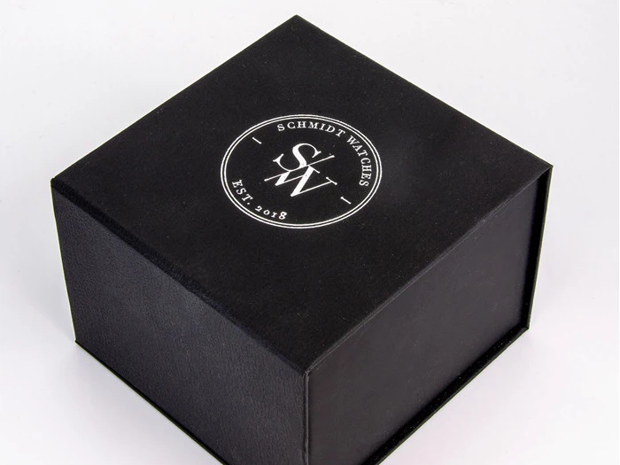 En sort æske med et rundt hvidt logo på toppen, hvor der står Schmidt Watches SW Est. 2018 på en almindelig hvid baggrund.