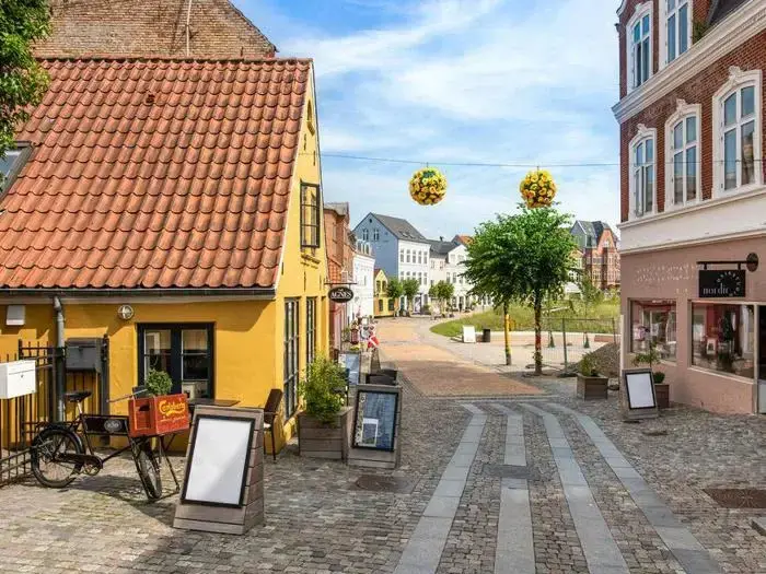 En brostensbelagt gade med gule og murede bygninger, udendørs skiltning, en cykel, træer og runde gule blomsterdekorationer, der hænger ovenover.