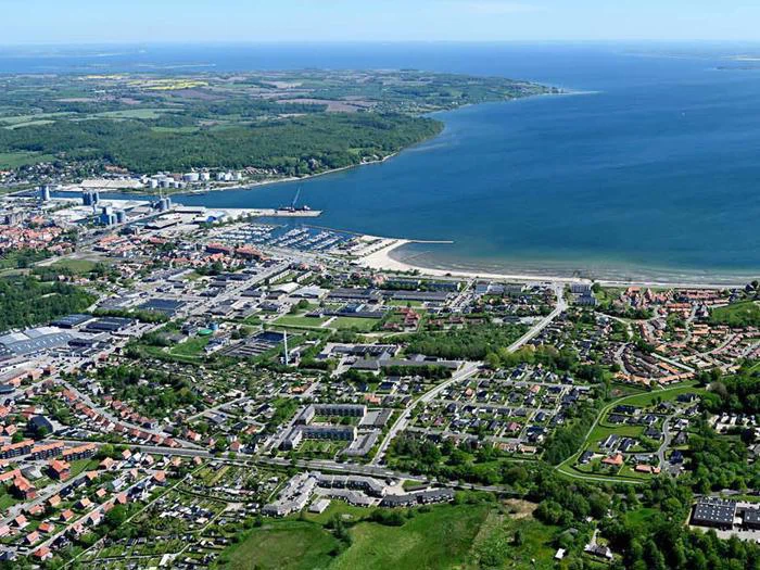 Luftfoto af en kystby med klynger af huse, marina, strand og omkringliggende grønne marker ved siden af en stor mængde blåt vand.