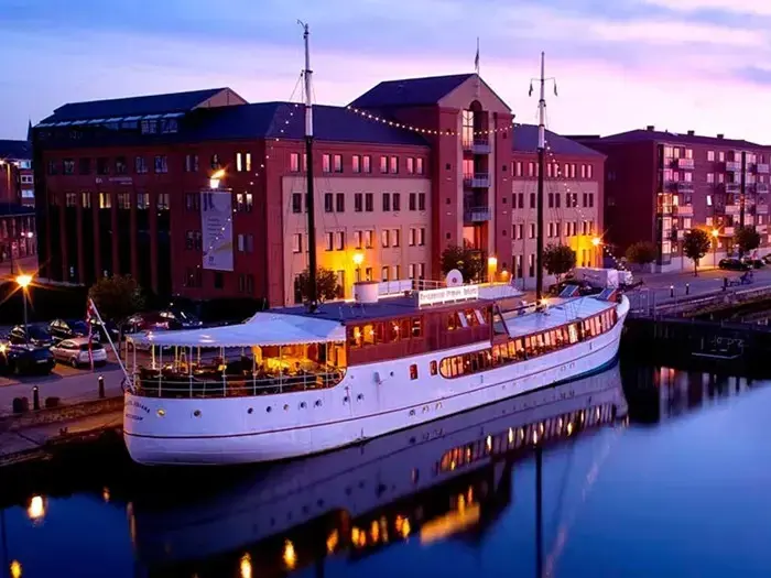 En stor hvid båd ligger ved siden af en moderne murstensbygning og spejler sig i det rolige vand i skumringen med blød, farverig himmelbelysning.