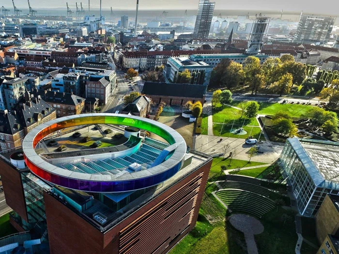 Luftfoto af en by med en bygning med en cirkulær, regnbuefarvet gangbro på taget omgivet af byrum og grønne områder.