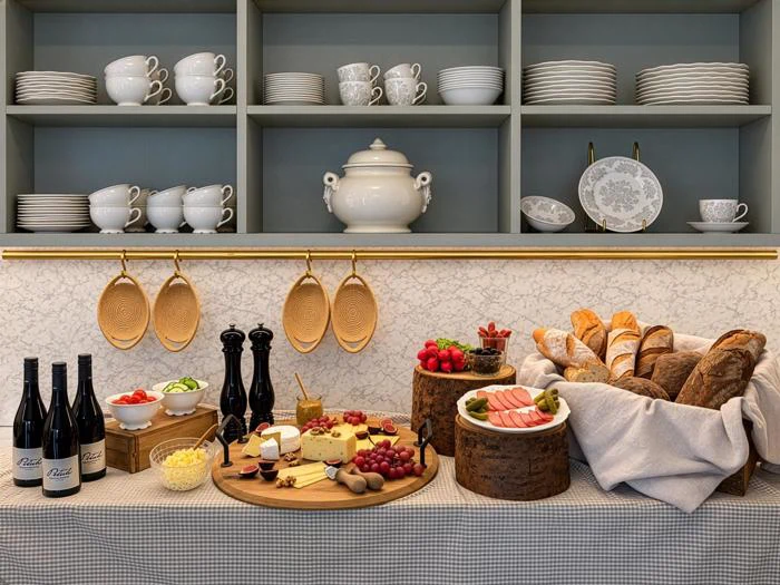 Et buffetbord med vin, ost, brød, pålæg, druer, frugt, tallerkener, kopper og en suppeterrin på hylderne, med en væg med marmormønster i baggrunden.