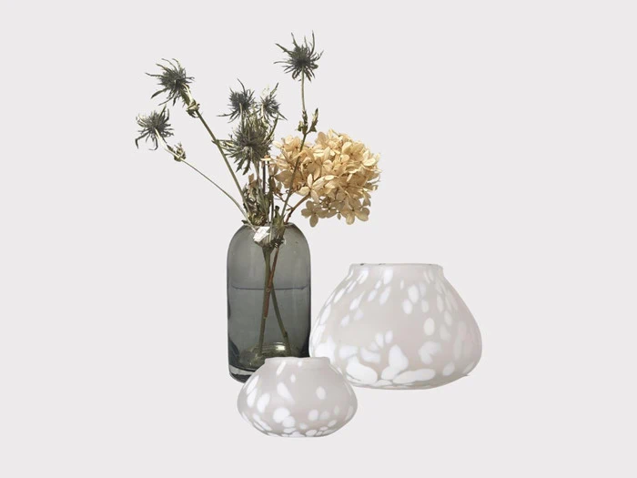 En glasvase med tørrede blomster står ved siden af to hvide, afrundede lysestager med bladmønster, alt sammen på en ensfarvet baggrund.