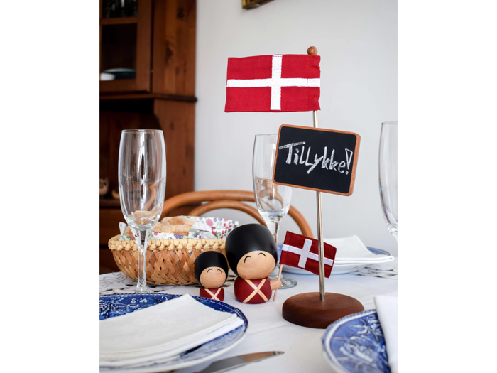 En borddækning med dansk tema med flag, soldaterfigurer i træ, en kurv med brød, vinglas og et kridttavleskilt med teksten Tillykke! omgivet af blå og hvide tallerkener.