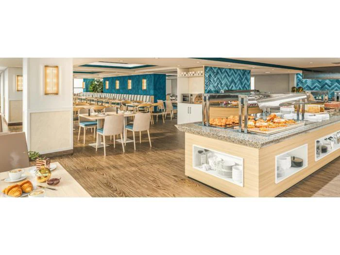 En lys, moderne buffetrestaurant med trægulve, blå detaljer, cremefarvede stole og en disk med forskellige kager og morgenmadsprodukter. I baggrunden er der dækket op til spisning.