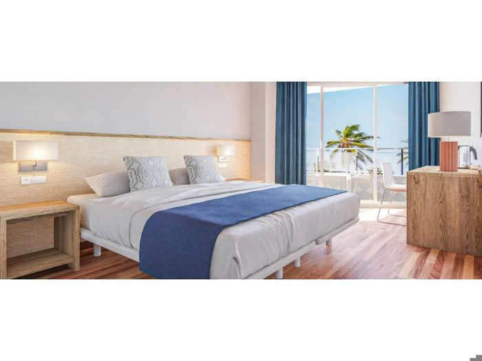 Et lyst hotelværelse med dobbeltseng, blåt tæppe og puder, træmøbler og en balkon med udsigt over havet og palmerne.