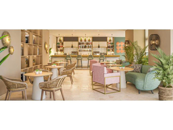 En moderne, stilfuld barlounge med kurvestole, pink og grønne sofaer, runde borde, planter og en velassorteret bar i baggrunden under pendellamper.