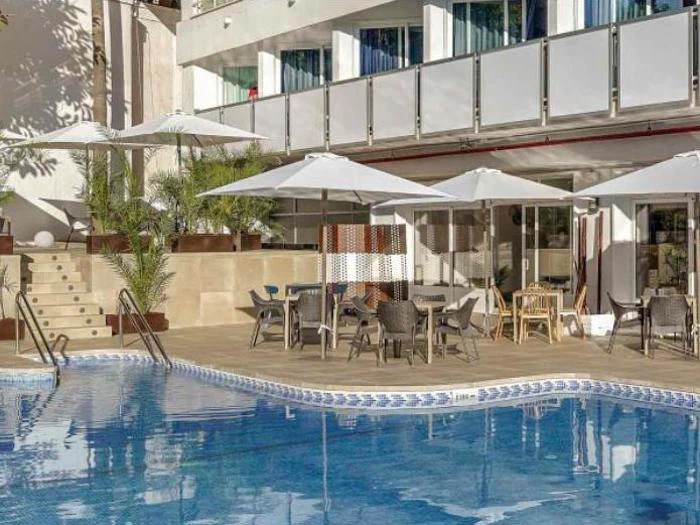 En swimmingpool med klart blåt vand, omgivet af liggestole, borde, parasoller og potteplanter ved siden af en moderne hotelbygning.