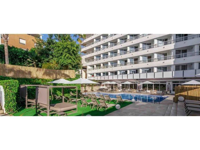 Udendørs udsigt over et moderne hotel med balkoner, liggestole ved poolen, solsenge, parasoller og en swimmingpool omgivet af grønne områder og træer.