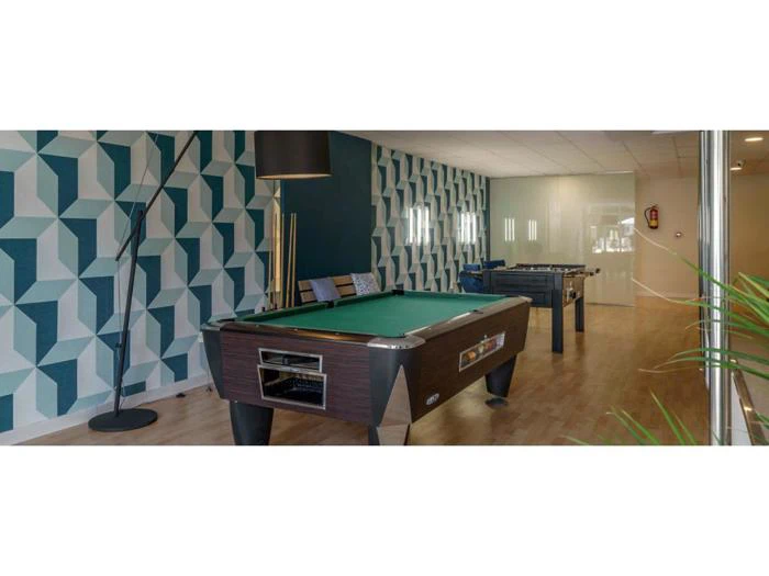 Et spillerum med et poolbord, et bordfodboldbord, en høj lampe, stole og geometrisk mønstret tapet på den ene væg.