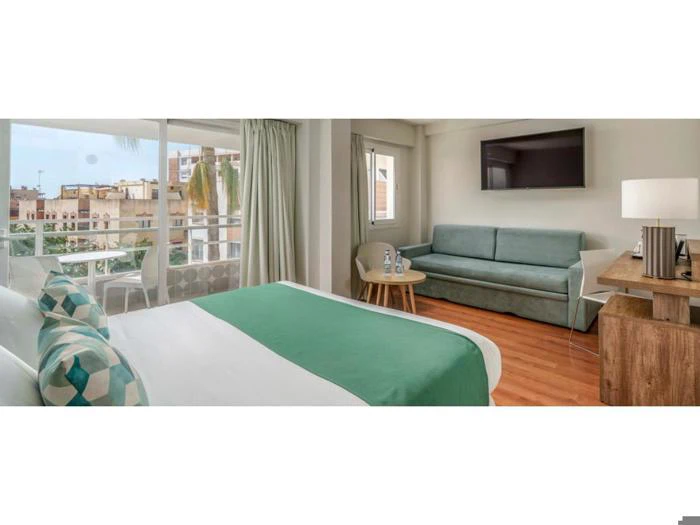 Et moderne hotelværelse med dobbeltseng, grønne detaljer, sofa, fladskærms-tv, store vinduer, et lille bord med vandflasker og et træskrivebord med lampe.