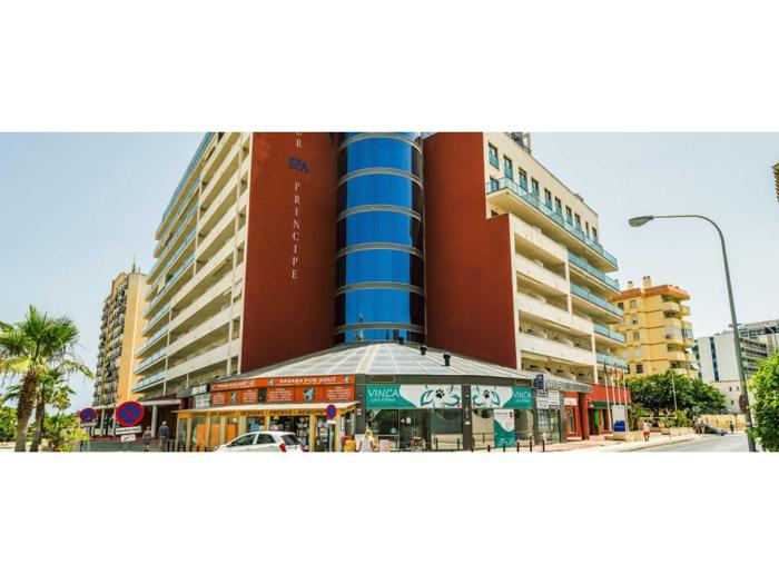 En moderne bygning i flere etager med balkoner, en afrundet sektion i blåt glas, butikker i stueetagen og palmer langs gaden på en solrig dag.