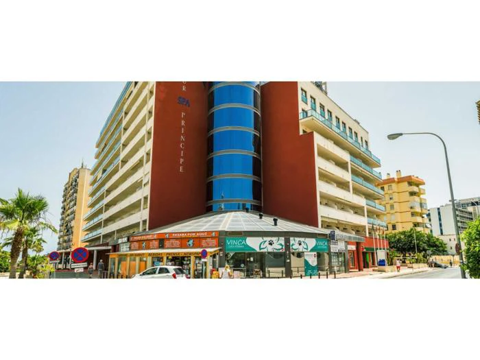 En moderne bygning i flere etager med balkoner, en afrundet sektion i blåt glas, butikker i stueetagen og palmer langs gaden på en solrig dag.