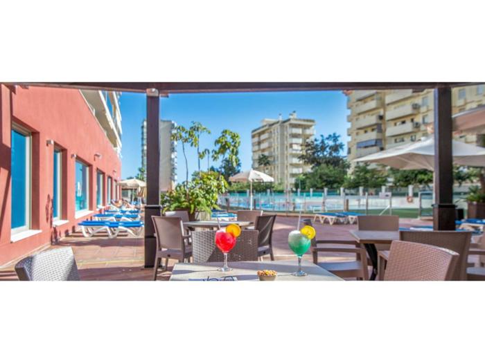 Udendørs siddeområde med drinks på et bord, liggestole ved en pool, parasoller og høje bygninger i baggrunden på en solrig dag.