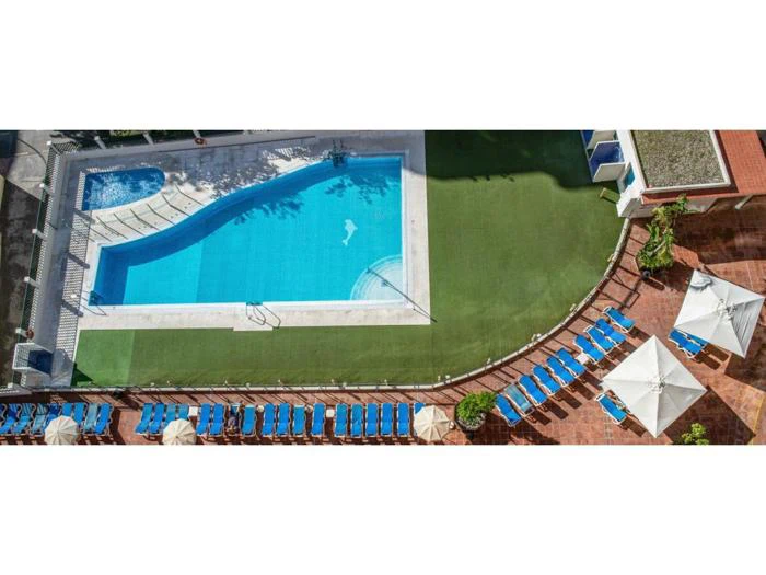 Luftfoto af en rektangulær swimmingpool med blå liggestole og hvide parasoller arrangeret på en terrasse og et grønt kunstgræsområde.