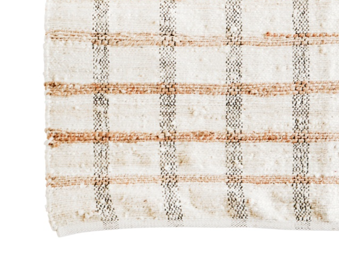 Et nærbillede af et beige vævet stof med et ternet mønster af brune og mørke linjer, der viser strukturerede fibre og let flossede kanter.