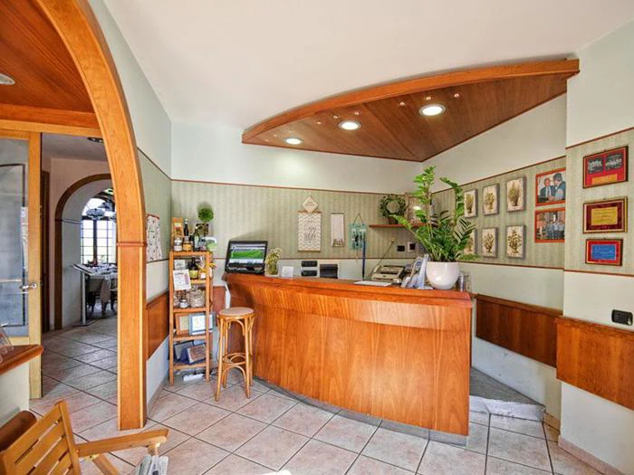 En lys hotelreception med et buet træskrivebord, certifikater på væggen, en computer, brochurer, en potteplante og en åben dør, der fører ind til et andet rum.