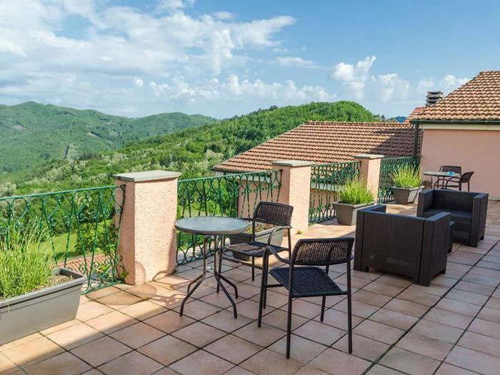 En solrig terrasse med kurvestole, et rundt bord, potteplanter og en smuk udsigt over grønne bakker og hustage under en blå himmel.