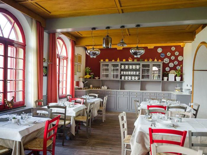 En hyggelig restaurant med hvide duge, træstole - nogle røde, andre hvide - store buede vinduer og en væg med hylder med fade og dekorative tallerkener.