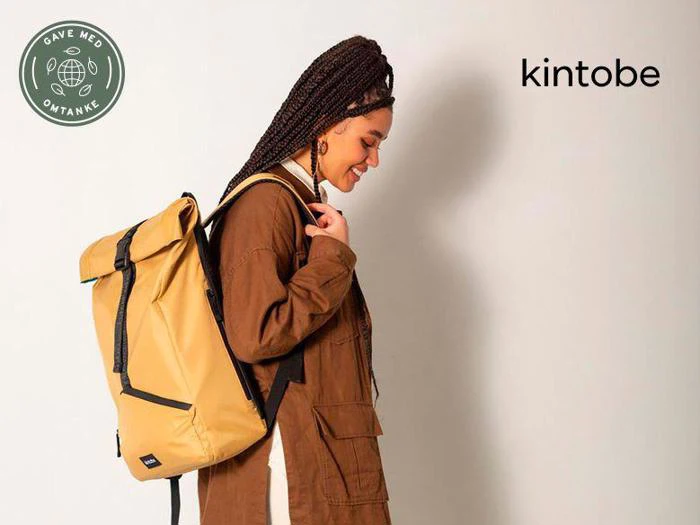 En kvinde med flettet hår smiler, mens hun bærer en stor gul rygsæk og er iført en brun jakke. Kintobe-logoet ses i øverste højre hjørne.