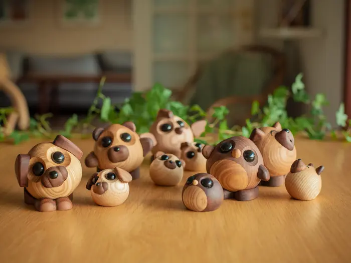 En gruppe små, runde dyrefigurer af træ med store, sorte øjne, anbragt på et træbord med grønne planter i baggrunden.
