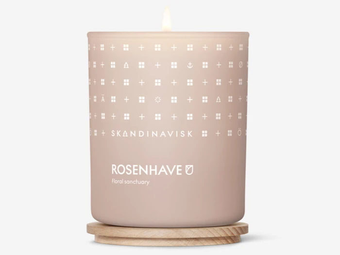 Et tændt beige lys mærket SKANDINAVISK ROSENHAVE Floral sanctuary står på en bordskåner af træ med et mønster af små hvide symboler på beholderen.