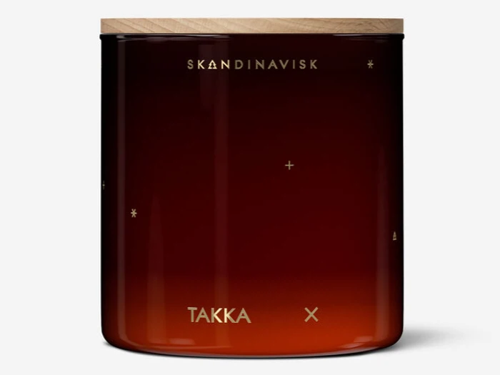 En mørk ravfarvet glaskrukke med trælåg og minimalistisk guldtekst med teksten SKANDINAVISK og TAKKA samt små guldstjerner og kors.