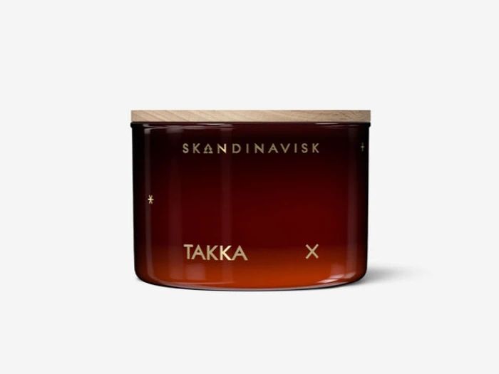 En lysestage af brunt glas med trælåg, mærket SKANDINAVISK og TAKKA med guldbogstaver på en almindelig hvid baggrund.
