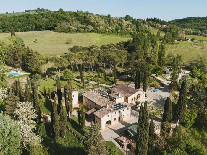 Luftfoto af en rustik villa omgivet af cypresser og grønne områder i et frodigt landskab med bakker og skov i baggrunden.