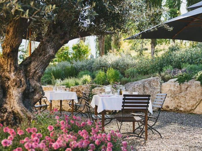 Udendørs restaurantborde med hvide duge mellem træer, blomster og grønne planter skaber en fredelig atmosfære i haven.