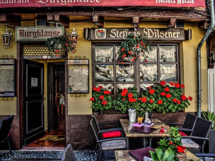 Udendørsservering på en rustik restaurant med røde blomster i altankasser, et menukort ved døren og borde på en brostensbelagt gade.